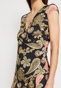 Femme portant une robe noire moulante avec des motifs floraux cachemire dorés, beiges et roses et un décolleté en V, montrant une partie de son torse et de son bras.
