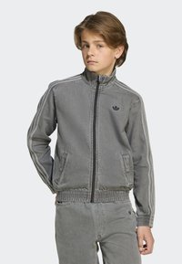 Veste en denim gris avec un col haut, des poches latérales, une taille élastique et un logo noir sur la poitrine. Présente des rayures blanches le long des manches.