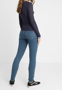 Marineblaues, geripptes Langarmoberteil kombiniert mit blauen Skinny-Jeans, die über Taschen und eine dezente Textur verfügen, ergänzt durch schwarze Sneaker.