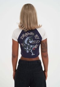 Camiseta recortada estilo béisbol con cuerpo en azul marino y mangas en crema. Presenta un gráfico de una pantera negra y el texto "DIBUJADO POR". Diseñada con una forma ajustada.