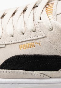 Beige mokkanahkakengät mustilla korostuksilla; varustettu valkoisilla nauhoilla ja kultaisella "PUMA"-logolla. Rakenne pintakuviointi näkyvissä olevilla saumoilla. Kumipohja.