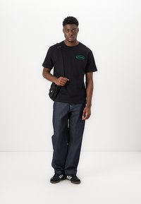 Svart t-skjorte med korte ermer og grønn logo, kombinert med mørkeblå baggy jeans og svarte joggesko med hvite striper. Bærer en liten svart veske.