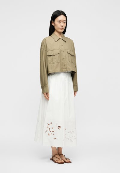 Γυναίκα με μακριά μαύρα μαλλιά στέκεται φορώντας ένα κομμένο χακί πράσινο πουκάμισο-μπουφάν με κουμπιά, λευκή φούστα με λουλουδάτες κοπές και καφέ σανδάλια.