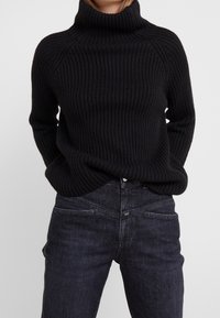 Jersey negro de cuello alto acanalado con un corte relajado y mangas largas, combinado con jeans vaqueros oscuros que cuentan con bolsillos delanteros y un lavado sutil.