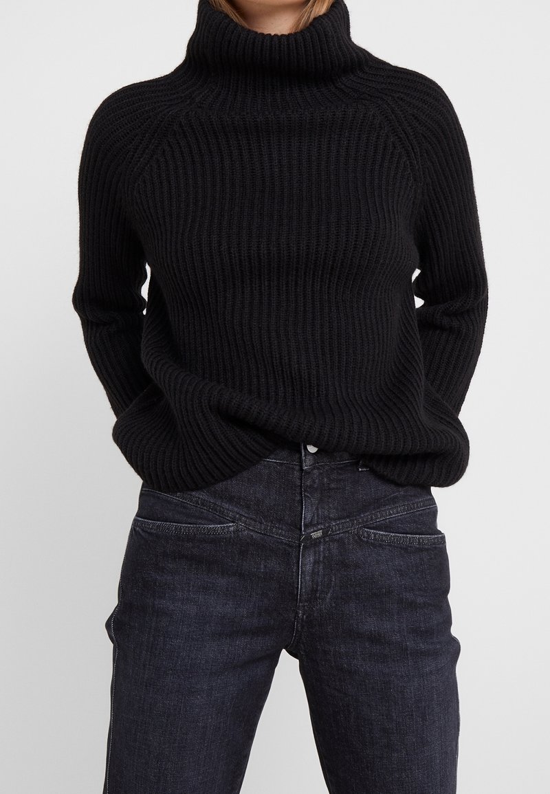 Jersey negro de cuello alto acanalado con un corte relajado y mangas largas, combinado con jeans vaqueros oscuros que cuentan con bolsillos delanteros y un lavado sutil.