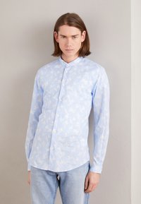 Camicia a maniche lunghe color blu chiaro con motivo floreale bianco, chiusura con bottoni, colletto a bandana e orlo dritto; abbinata a jeans in denim chiaro.