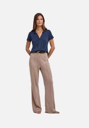 Donna che indossa una camicia blu navy a maniche corte con bottoni, pantaloni beige a gamba larga, cintura marrone, collana d'oro e sandali aperti, in piedi con le mani dietro.