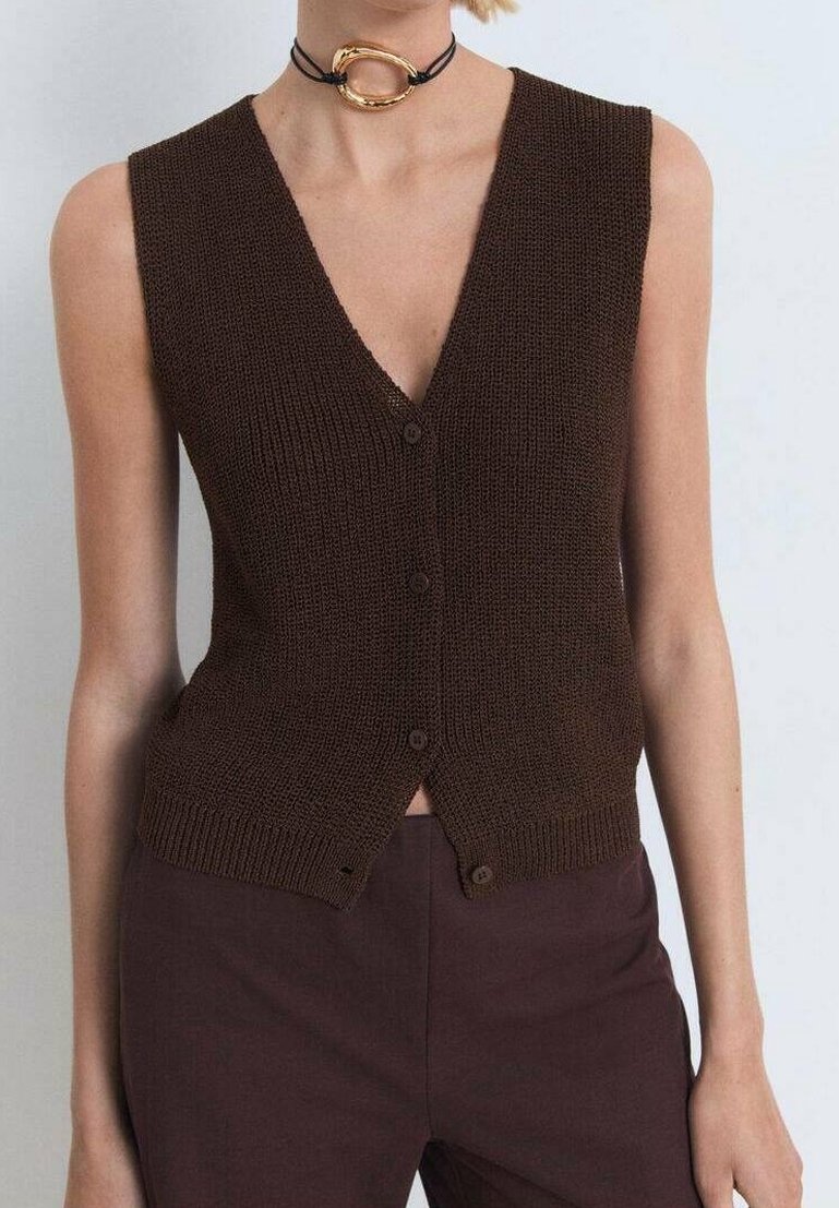 Brązowa, dzianinowa, bezrękawnik z głębokim dekoltem w kształcie litery V, z zapięciem na guziki i prążkowanym dołem. Noszony na ciemnych brązowych spodniach, z złotą naszyjnikiem typu choker.