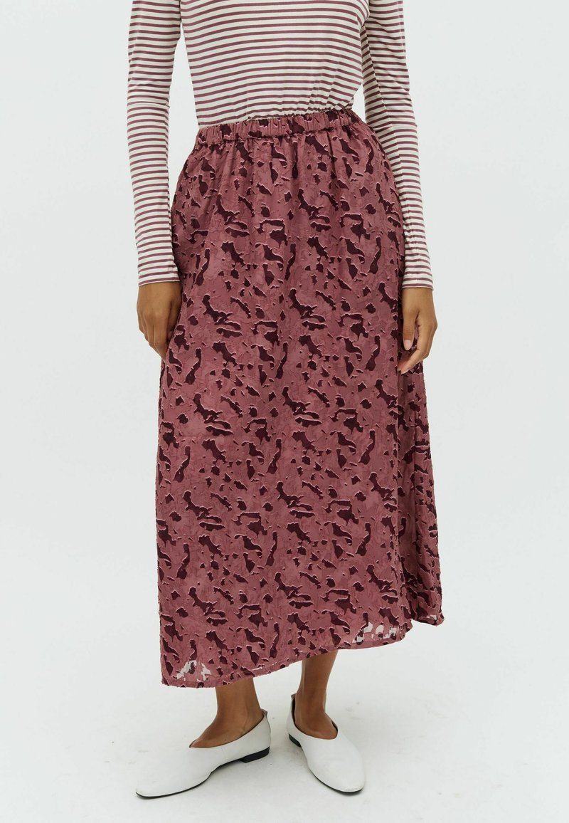 Jupe maxi en rose poussiéreux avec un motif texturé et abstrait. Taille élastique, tissu fluide, associée à des chaussures blanches à enfiler.