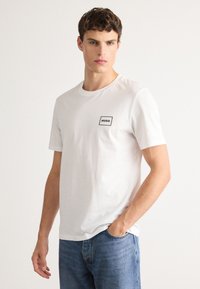 Ung mann som har på seg en hvit T-skjorte med korte ermer og et lite "HUGO"-logo på brystet, sammen med blå jeans, står mot en ensfarget bakgrunn.