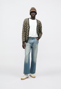 Καφέ και μπεζ καρό ζακέτα πάνω από λευκό μπλουζάκι, συνδυασμένη με ανοιχτό μπλε ξεβαμμένο τζιν και λευκά αθλητικά παπούτσια με σόλα από καουτσούκ. Πολύχρωμο πλεκτό καπέλο.