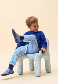 Kind, der auf einem blauen, gepunkteten Stuhl sitzt, trägt einen blauen Sweatshirt und helle blaue Hosen mit bunten Streifen sowie gemusterte blaue Schuhe.