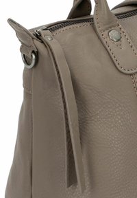 Primo piano di una cerniera di una borsa in pelle taupe con tirante, cuciture visibili, rivetto in metallo e anello di attacco del manico.