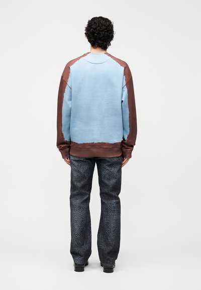 Άτομο με σγουρά μαλλιά που φορά μπλε και καφέ μακρυμάνικη μπλούζα και σκούρο παντελόνι με σχέδια, στέκεται με την πλάτη γυρισμένη σε απλό φόντο.