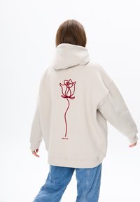 Beige hoodie met een rozenontwerp in donkerrood geborduurd op de rug. Heeft een relaxte pasvorm en een capuchon met trekkoord.