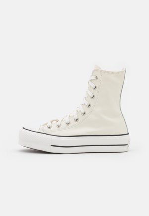 Baskets montantes blanches en toile avec lacets blancs, semelle en caoutchouc avec double rayures noires et œillets métalliques sur fond blanc uni.