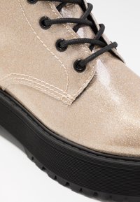 Close-up van een glinsterende beige veterlaars met zwarte veteroogjes en een dikke zwarte geribbelde zool.