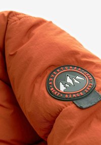 Tissu orange à finition texturée, avec un patch logo circulaire noir orné d'accents rouges et de graphiques de montagnes, fixé avec une sangle grise.