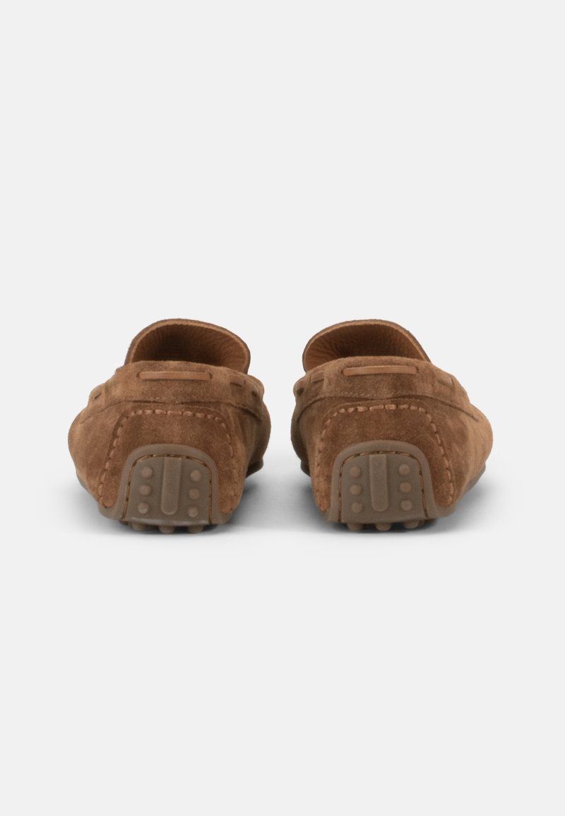 Een paar bruine suede loafers met rubberen hielgrepen, gezien van achteren op een lichtgrijze achtergrond.