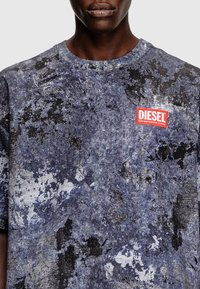 Muž v modro-černém tričku Diesel s vzorem a červenou nášivkou loga na hrudi, umístěný na jednobarevném pozadí.