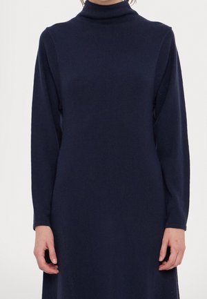 Personne portant une robe col roulé bleu marine à manches longues, montrée du cou jusqu'à mi-cuisse, les mains détendues sur les côtés.