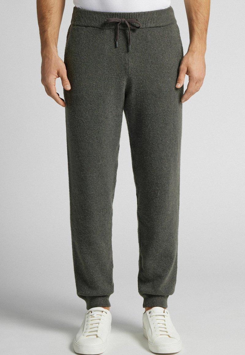 Šedé sametové joggingové kalhoty s elastickým pasem a šňůrkou. Hladké manžety a boční kapsy zvyšují funkčnost. V páru s bílými teniskami.