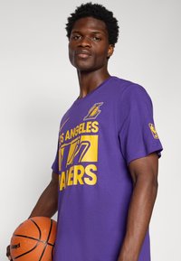 Purpurinė medvilninė marškinėliai su geltona "Los Angeles Lakers" grafika. Trumpomis rankovėmis, apvaliu kaklu, dėvi žmogus, laikantis krepšinį.