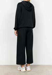 Černá mikina s kapucí na šňůrku, v kombinaci s volnými černými kalhotami. Hladká tkanina, pohodlný střih a nošeno s světlými teniskami.