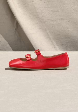 Rdeč usnjen čevelj Mary Jane z dvema zlatima zaponkama na svetlo bež površini in tkanem ozadju.