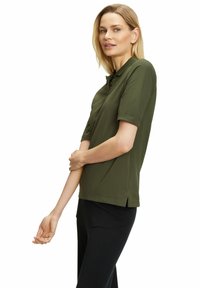 Olijfgroene polo met korte mouwen, drie knoopsluitingen, zijsplitten aan de zoom en een gladde kledingstructuur. Zwarte broek.