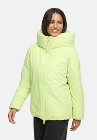 Vrouw die glimlacht in een lichtgroene pufferjack met hoge kraag en zwarte broek, staand tegen een effen lichte achtergrond.