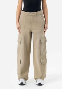 Beige cargo bukser med brede ben, laget av slitesterkt stoff. Har flere store lommer, knapping og synlige sømmer.