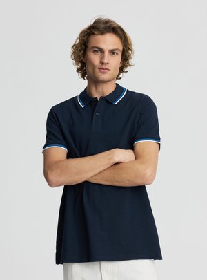 Giovane uomo con capelli ricci che indossa una polo blu navy con colletto e polsini a righe bianche e blu, in piedi con le braccia incrociate.