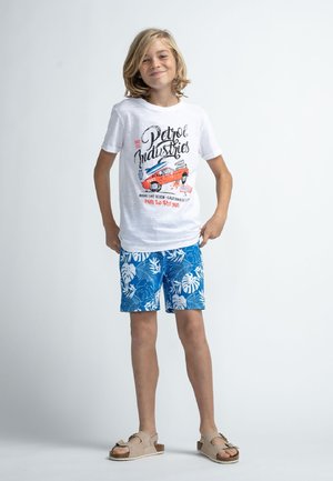 Camiseta blanca con estampado de "Petrol Industries" y gráfico de un coche rojo; pantalones cortos azules con patrón de hojas tropicales blancas; sandalias beige.