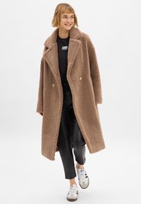 Doppelreihiger brauner, wuscheliger Mantel mit breitem Kragen, weicher Textur und großen Knöpfen, kombiniert mit schwarzen Lederhosen und weißen Turnschuhen.