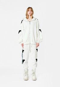 Sudadera blanca con cremallera y capucha, con diseños bordados de alas negras, combinada con pantalones deportivos a juego que presentan acentos similares. Estilizado con botas blancas.