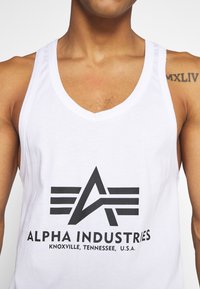 Bílý tílko s černým grafickým logem s úhlovým designem a textem, který zní "ALPHA INDUSTRIES" a "KNOXVILLE, TENNESSEE, U.S.A."