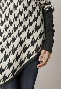 Zwart en crèmekleurig gemarmerde gebreide poncho met driehoekig ontwerp, grijze geribbelde mouwen en een klein bruin logo detail nabij de onderrand.