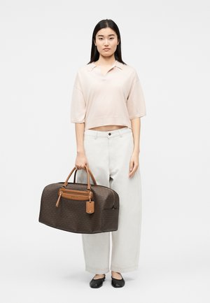 Naine beežikat värvi lühikese särgi ja valgete pükstega, hoiab suurt pruuni nahast käekotti, millel on nahatoonides sangad ja detailid.