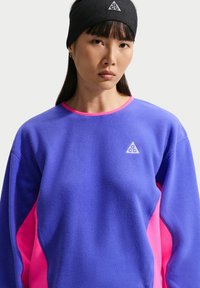 Γυναίκα που φοράει μπλε και ροζ φλις μπλούζα με λευκό τριγωνικό λογότυπο και μαύρη κορδέλα με ταιριαστό λογότυπο.