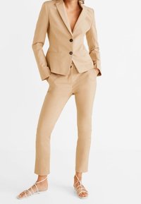 Lichtbeige maatpak met een getailleerde blazer en slanke broek, voorzien van twee knopen aan de voorkant en een zachte stof. Draag het met sandaaltjes met bandjes.