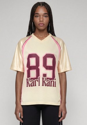 Jeune femme aux cheveux tressés portant un maillot de sport beige et rose avec le numéro 89 et le texte "Karl Kani" à l'avant.
