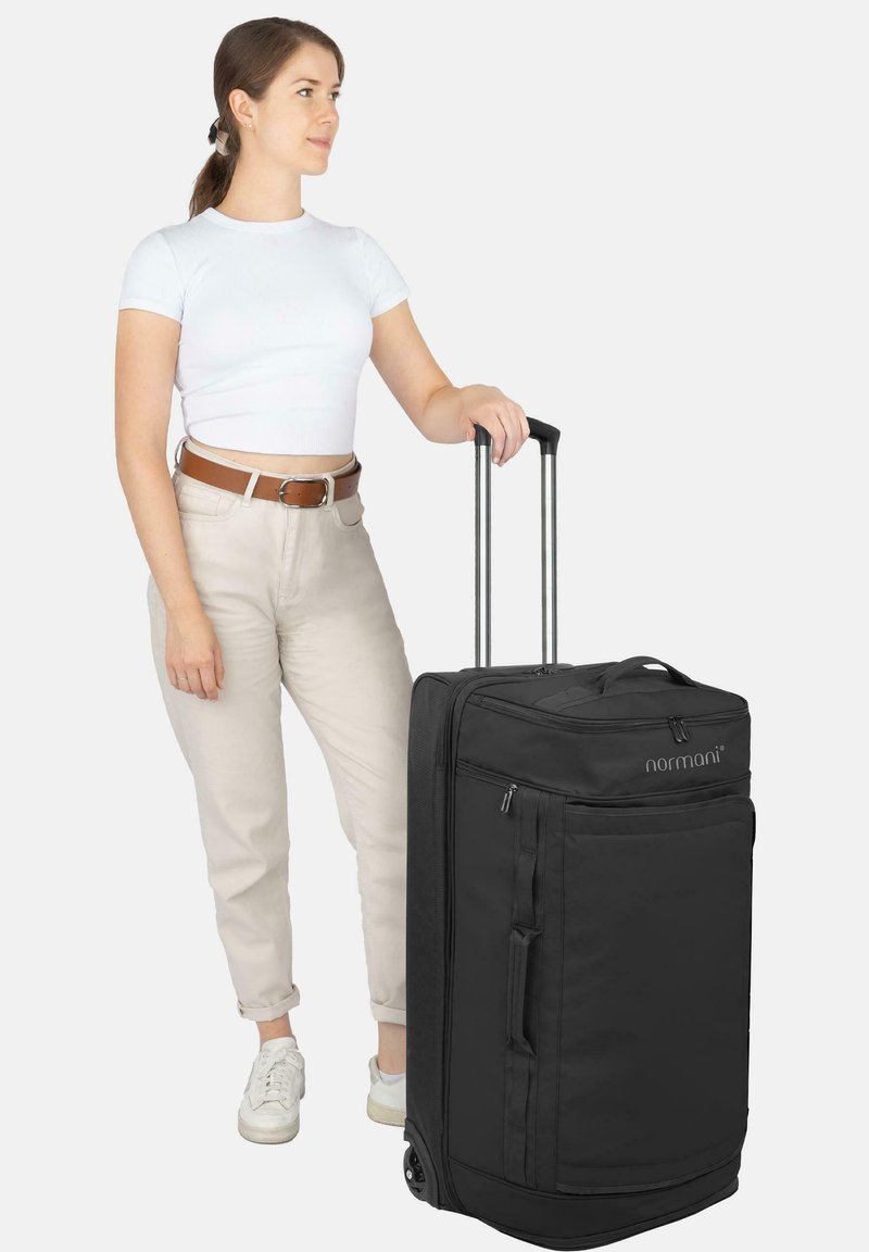 Schwarzer Stoffkoffer mit zwei Rädern, Teleskopgriff, Seitentasche und Reißverschluss, neben einer Person in heller Kleidung platziert.