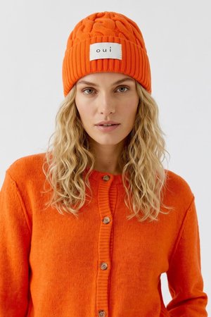 Blonde Frau mit welligem Haar trägt eine orange gestrickte Mütze mit "oui"-Label und eine passende orangefarbene, geknöpfte Strickjacke.