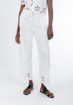 Weißes Leinenoutfit mit einem kurzen Oberteil mit Blumen-Aussparungen und passenden weiten Hosen mit ähnlichen Blumenmustern am Saum. Schwarze Sandalen.