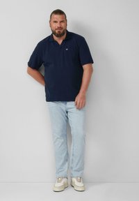 Granatowa koszulka polo z krótkim rękawem i kieszenią, zestawiona z jasnoniebieskimi prostymi dżinsami oraz kremowymi sneakersami, stojąca na szarym tle.