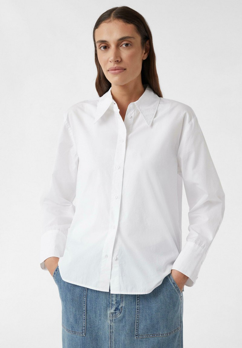 Woman with long brown hair wearing a white button-up shirt with wide collar and blue denim skirt, hands in pockets, neutral background.