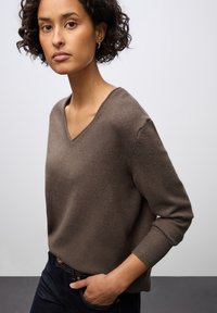 Hnědý svetr s výstřihem do V a dlouhými rukávy, vyrobený z měkké pleteniny. Má žebrované manžety a volný střih, zkombinovaný s tmavými džíny.