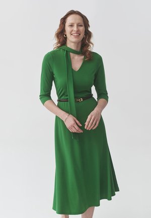 Robe verte avec un décolleté en V, des manches longues et un accent à nouer autour du cou. Taille ajustée avec une ceinture marron. Tissu lisse et fluide.