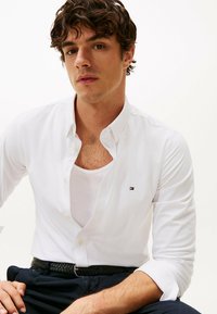 Camicia bianca con colletto classico, dotata di un piccolo logo sul petto, indossata sopra una canotta bianca, abbinata a pantaloni scuri.